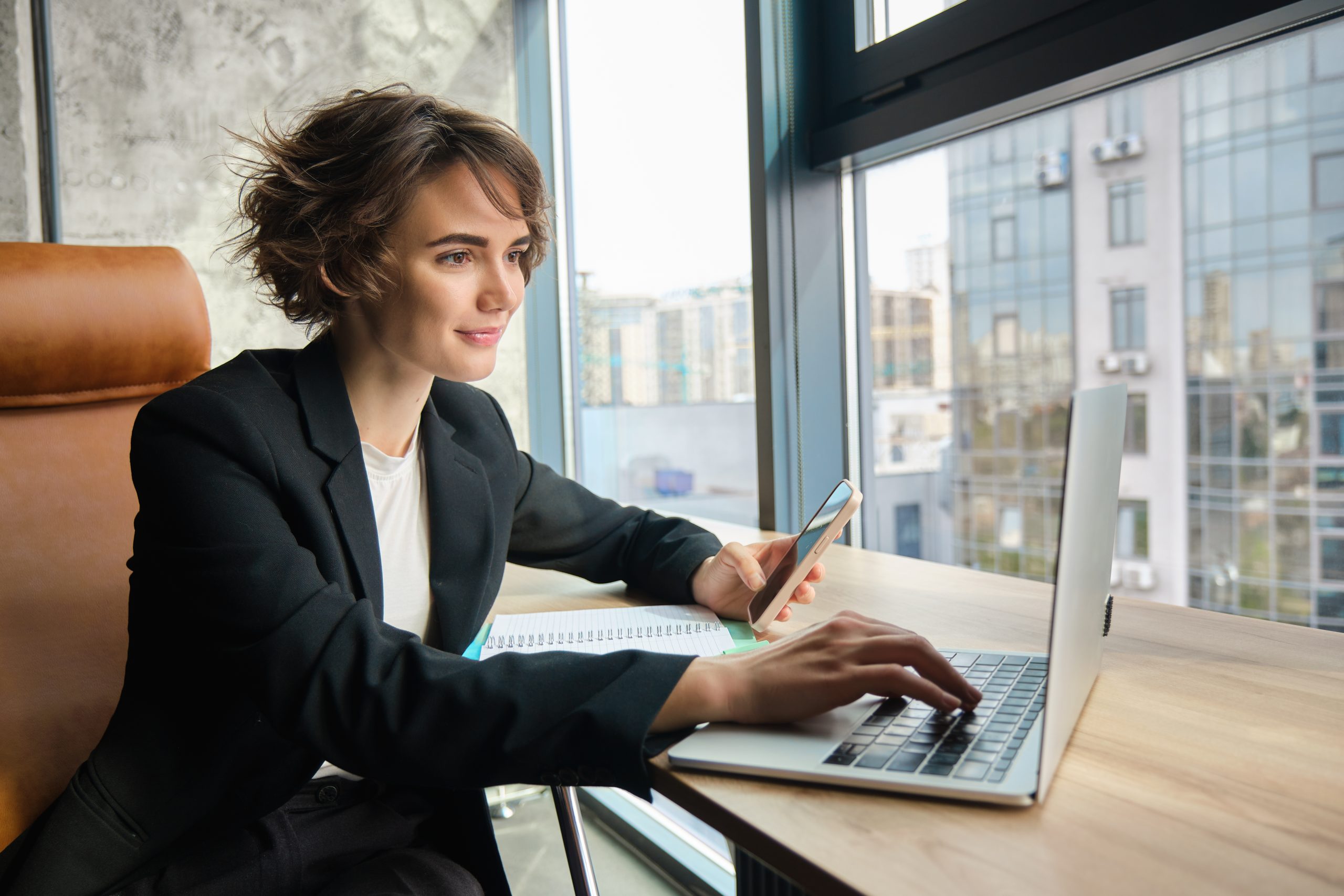Portrait of businesswoman working on laptop, using mobile phone, sitting in an office, looking through her documents and workload, sharing coworking space